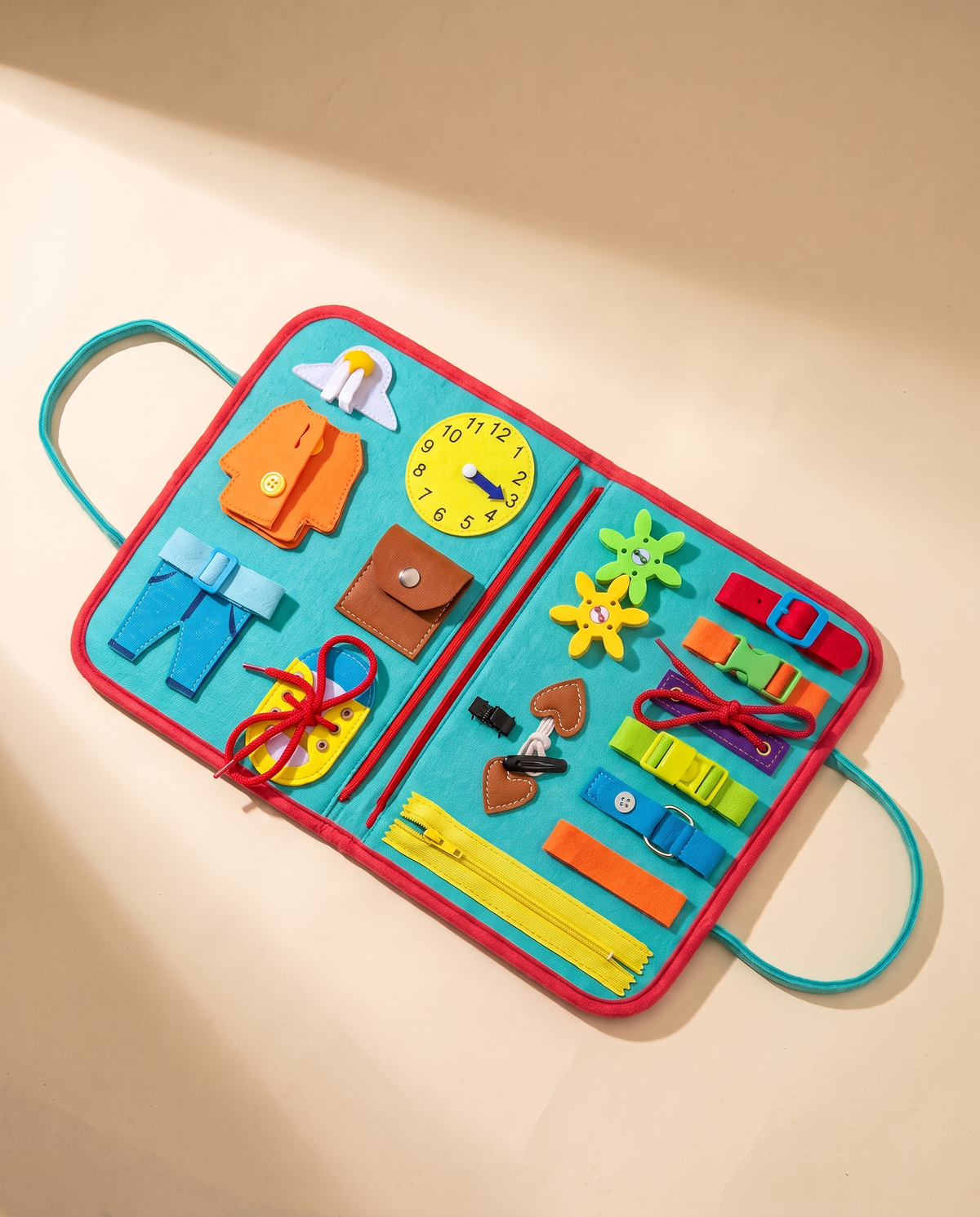 Open turquoise busy book with red trim and carry handles, showing zips, buckles, laces, buttons and a toy clock, on a cream background