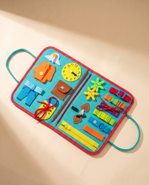 Open turquoise busy book with red trim and carry handles, showing zips, buckles, laces, buttons and a toy clock, on a cream background