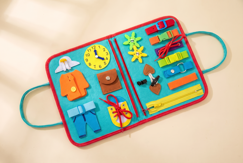 Turquoise busy book laid open flat with a yellow zip, coloured buckles and straps, laces and a toy clock